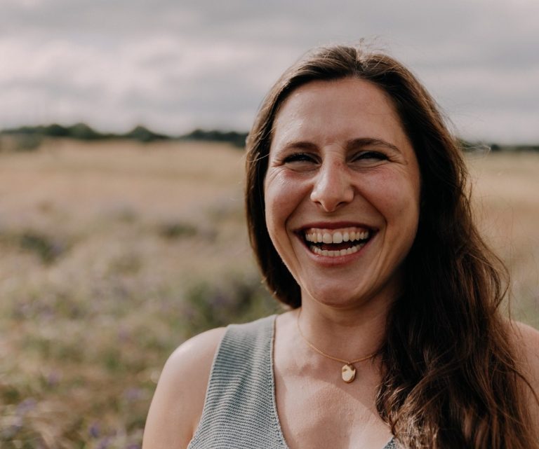 Lachende Frau mit langen Haaren in einem Feld unter bewölktem Himmel.